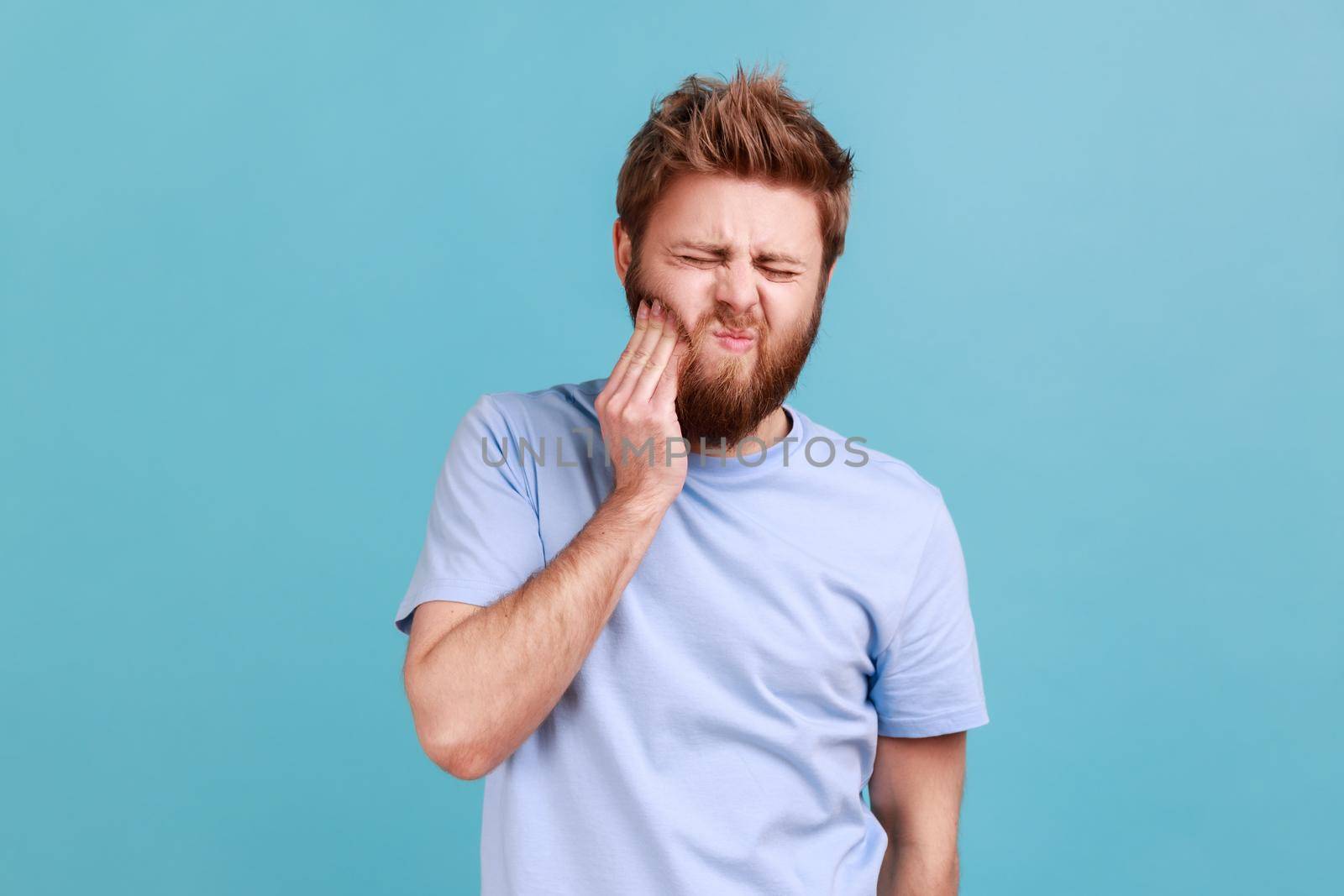 Portrait of unhealthy sick bearded man pressing sore cheek, suffering acute toothache, periodontal disease, cavities or jaw pain. Indoor studio shot isolated on blue background.