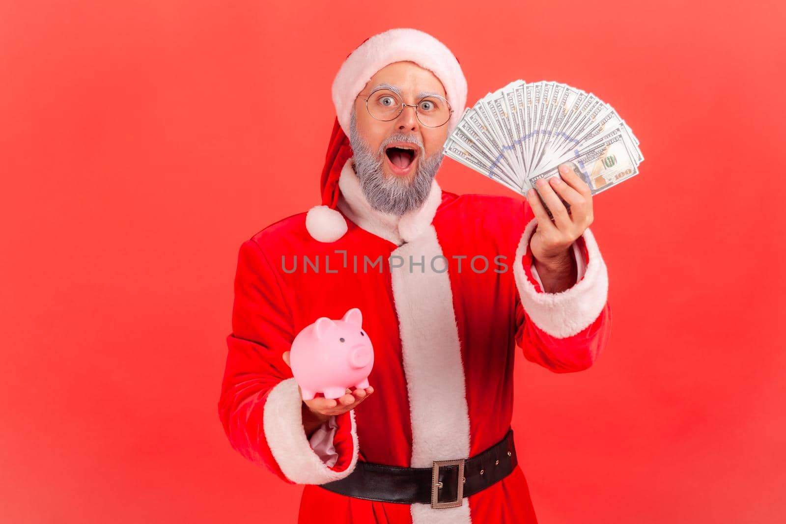 Surprised elderly man with gray beard wearing santa claus costume holding dollars banknotes and piggy bank, savings, high rate deposit. Indoor studio shot isolated on red background.