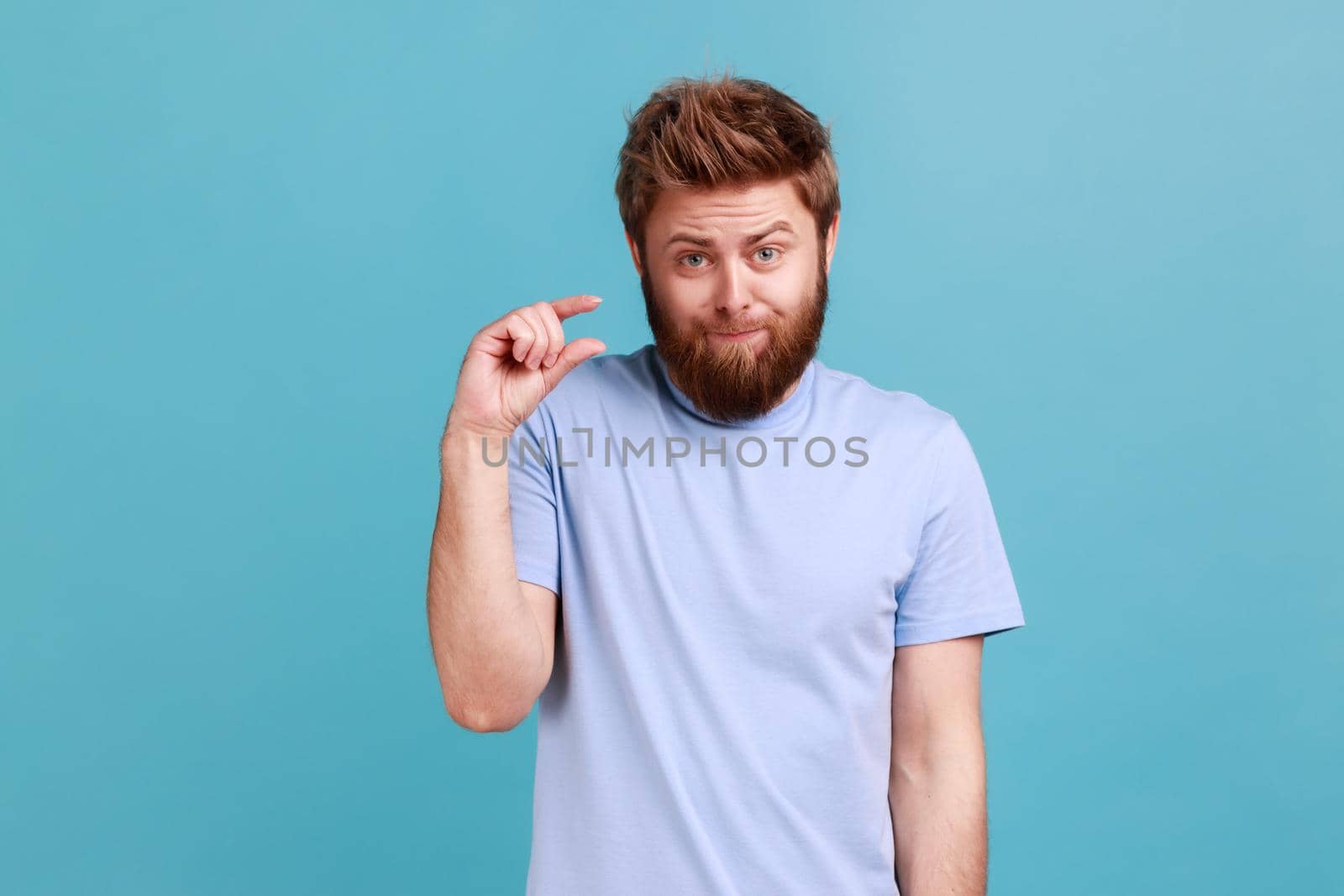 Portrait of disappointed bearded man showing a little bit gesture, dissatisfied with low rating, measuring scale, frowning face and looking at camera. Indoor studio shot isolated on blue background.