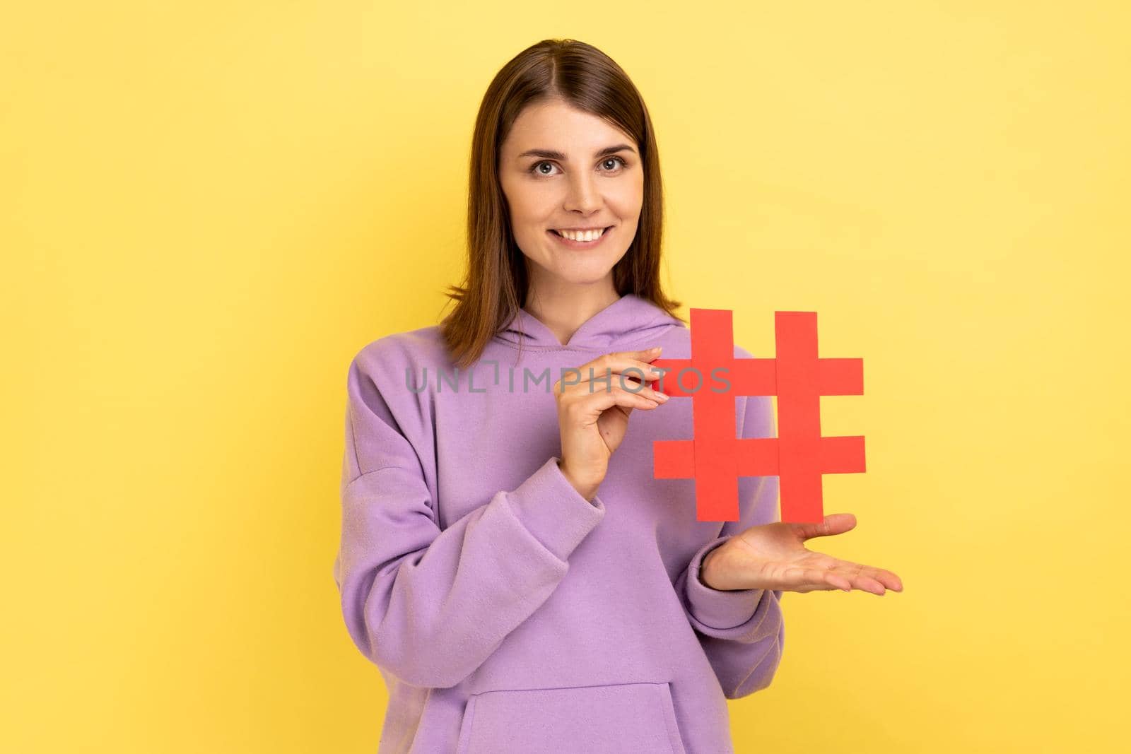 Portrait of young woman standing presenting red hashtag, tagging blog trends, viral topic in social network, wearing purple hoodie. Indoor studio shot isolated on yellow background.
