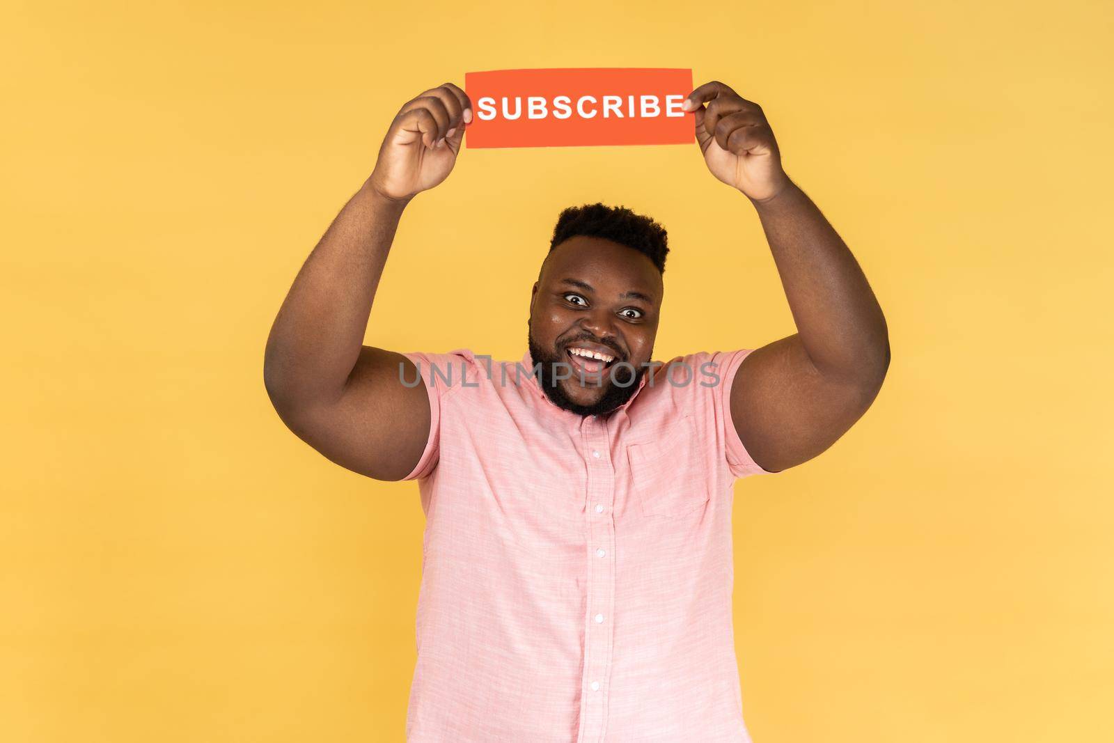 Portrait of excited amazed man in pink shirt holding with paper card with subscribe inscription above head, looking at camera with toothy smile. Indoor studio shot isolated on yellow background.