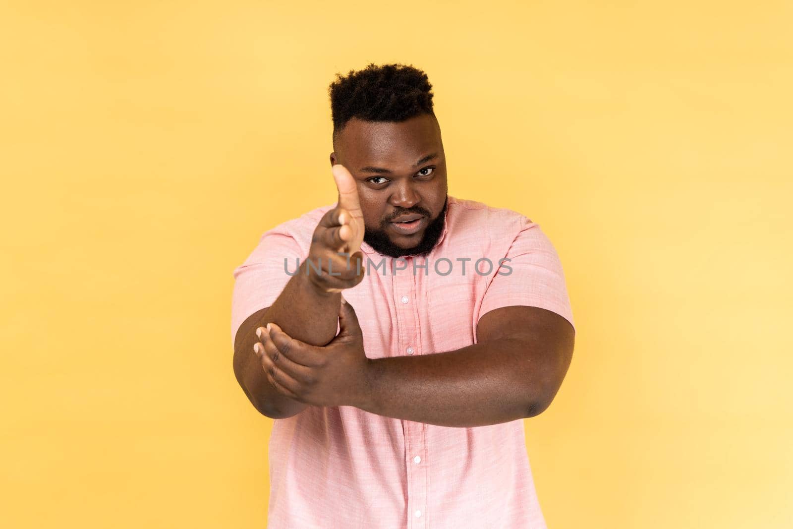 I'll kill you. Portrait of man wearing pink shirt pointing finger guns to camera and looking aggressive, threatening to shoot, hands imitating weapon. Indoor studio shot isolated on yellow background.
