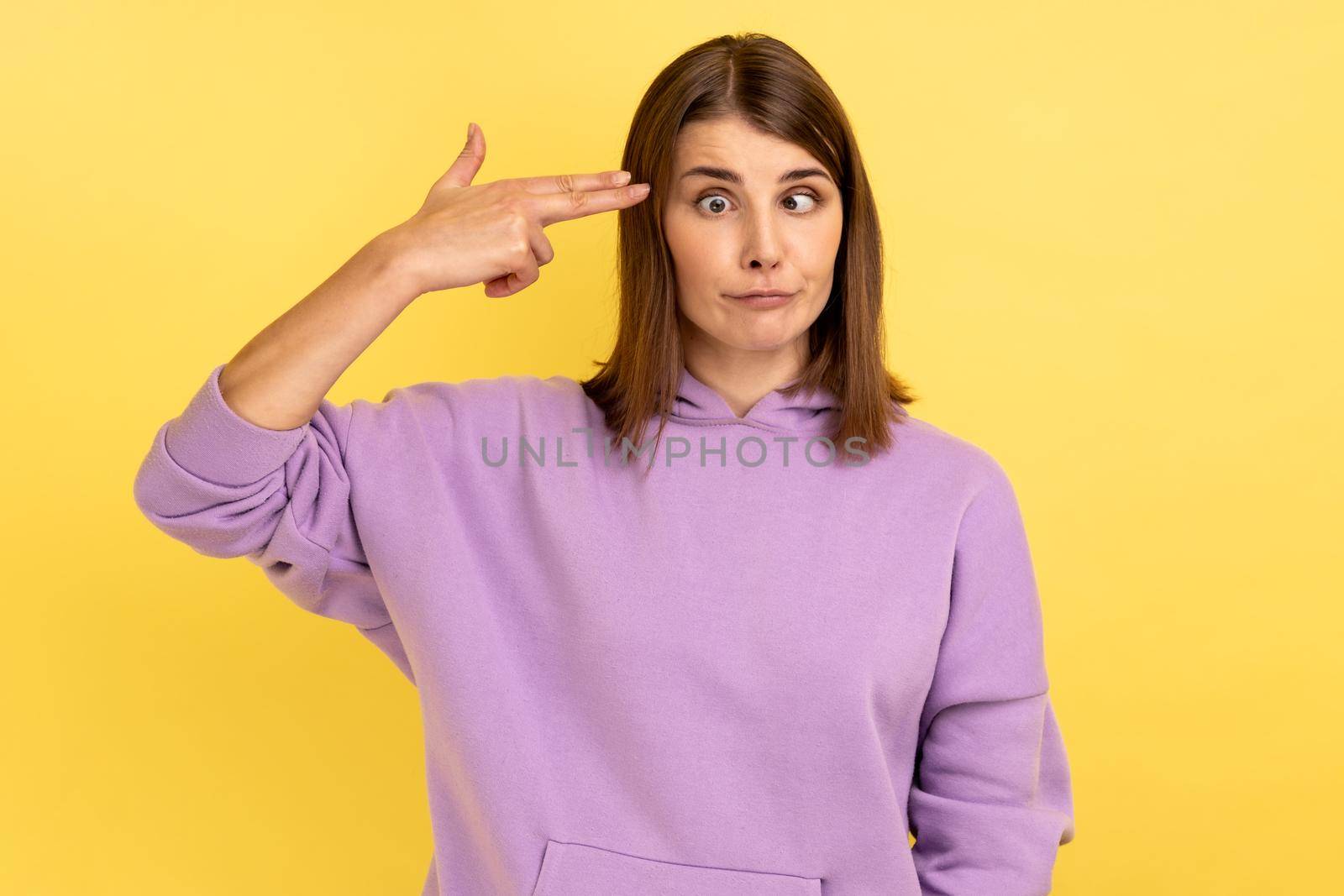 Portrait of desperate woman, pointing finger gun to head, shooting herself, making suicide gesture to stop depression, wearing purple hoodie. Indoor studio shot isolated on yellow background.