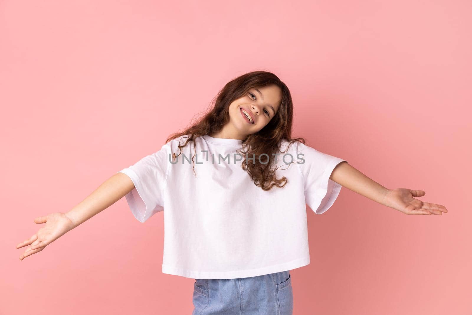 Portrait of positive optimistic little girl wearing white T-shirt inviting to embrace, keeping hands as giving, sharing for free. Indoor studio shot isolated on pink background.
