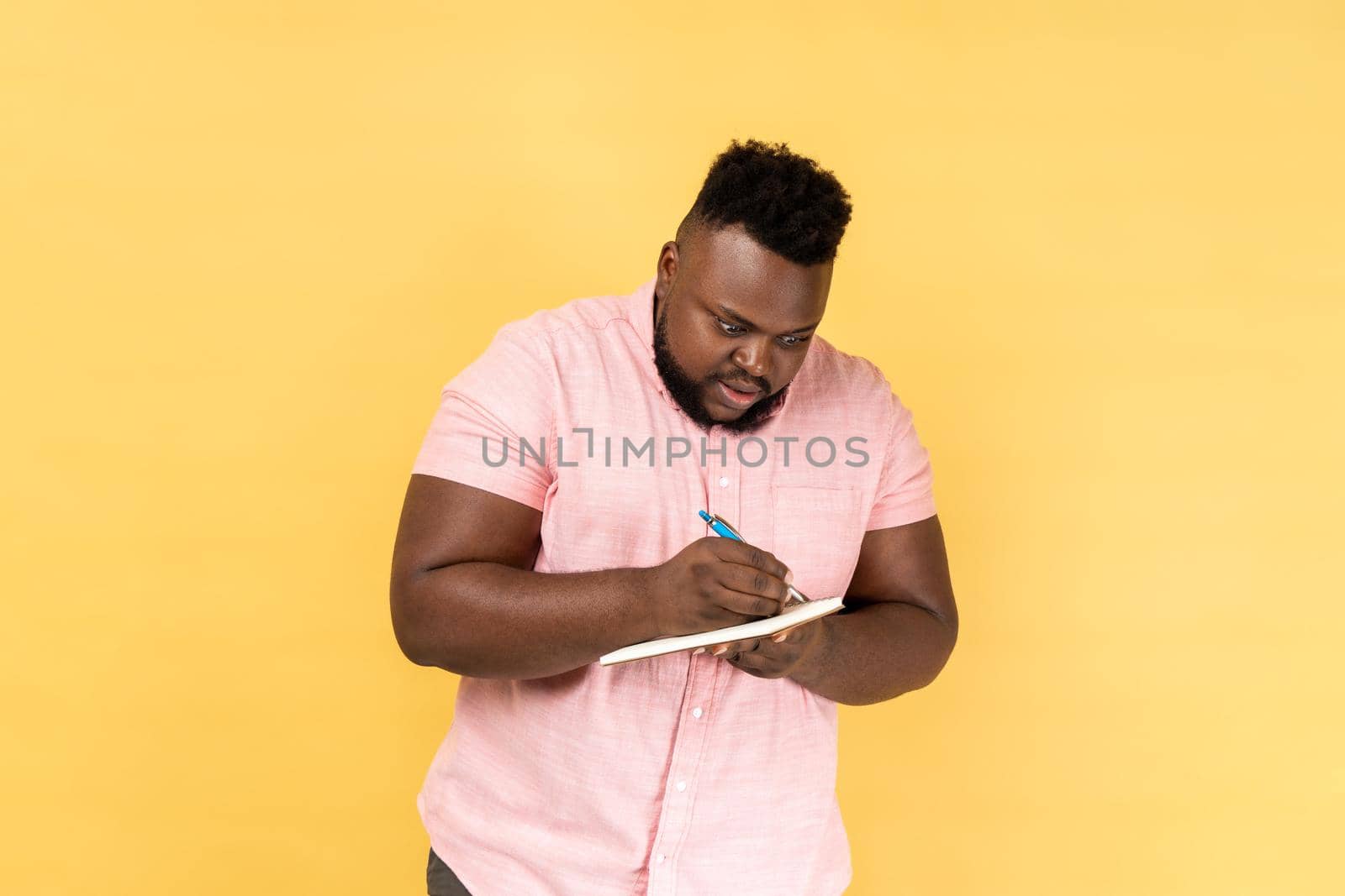 Portrait of astonished man wearing pink shirt writing in notebook, completing to-do list or studying, having surprised expression. Indoor studio shot isolated on yellow background.