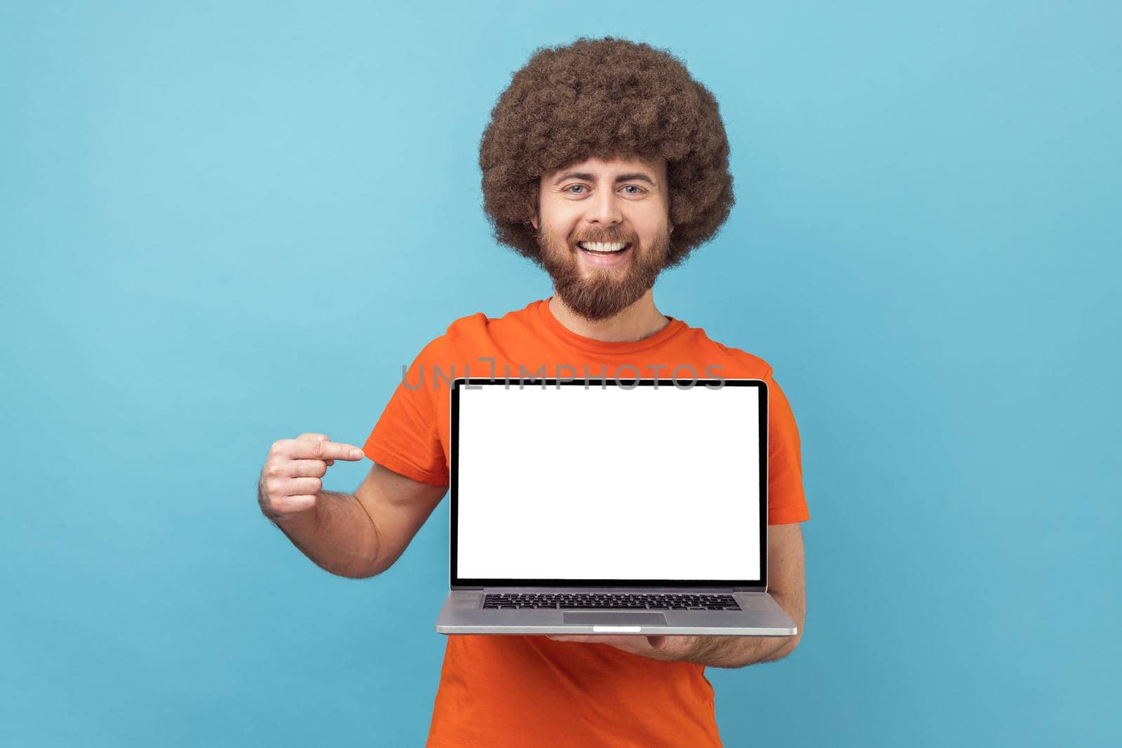 Portrait of delighted man with Afro hairstyle wearing orange T-shirt standing showing empty display of laptop, pointing at screen, advertising area. Indoor studio shot isolated on blue background.