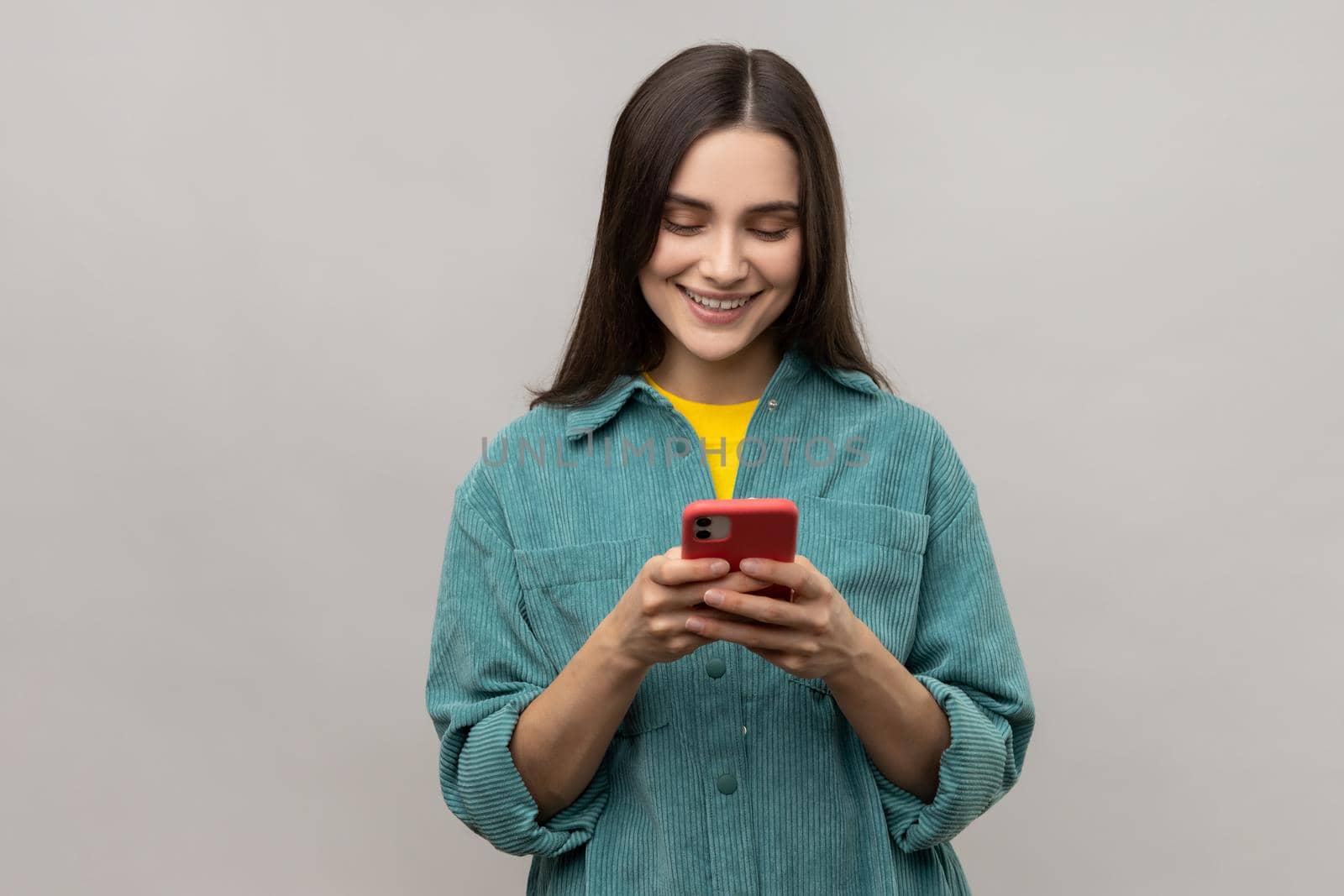 Happy pretty woman reading message on smartphone and smiling, using mobile device for communication, browsing web, wearing casual style jacket. Indoor studio shot isolated on gray background.