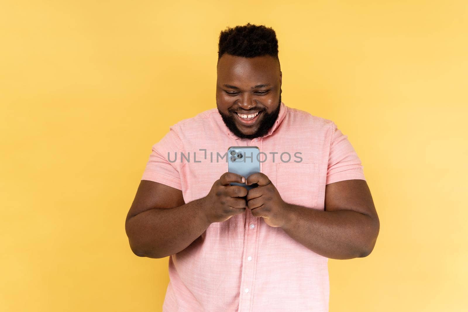 Portrait of bearded happy man wearing pink shirt using mobile phone with happy expression, addicted to smartphone, texting in social network. Indoor studio shot isolated on yellow background.