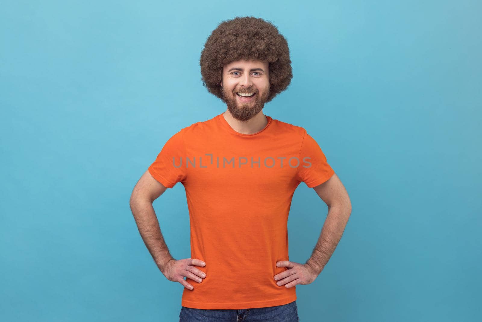 Portrait of cheerful man wearing orange T-shirt looking at camera with toothy smile and happy facial expression, being in good mood, keeps hands on hips. Indoor studio shot isolated on blue background