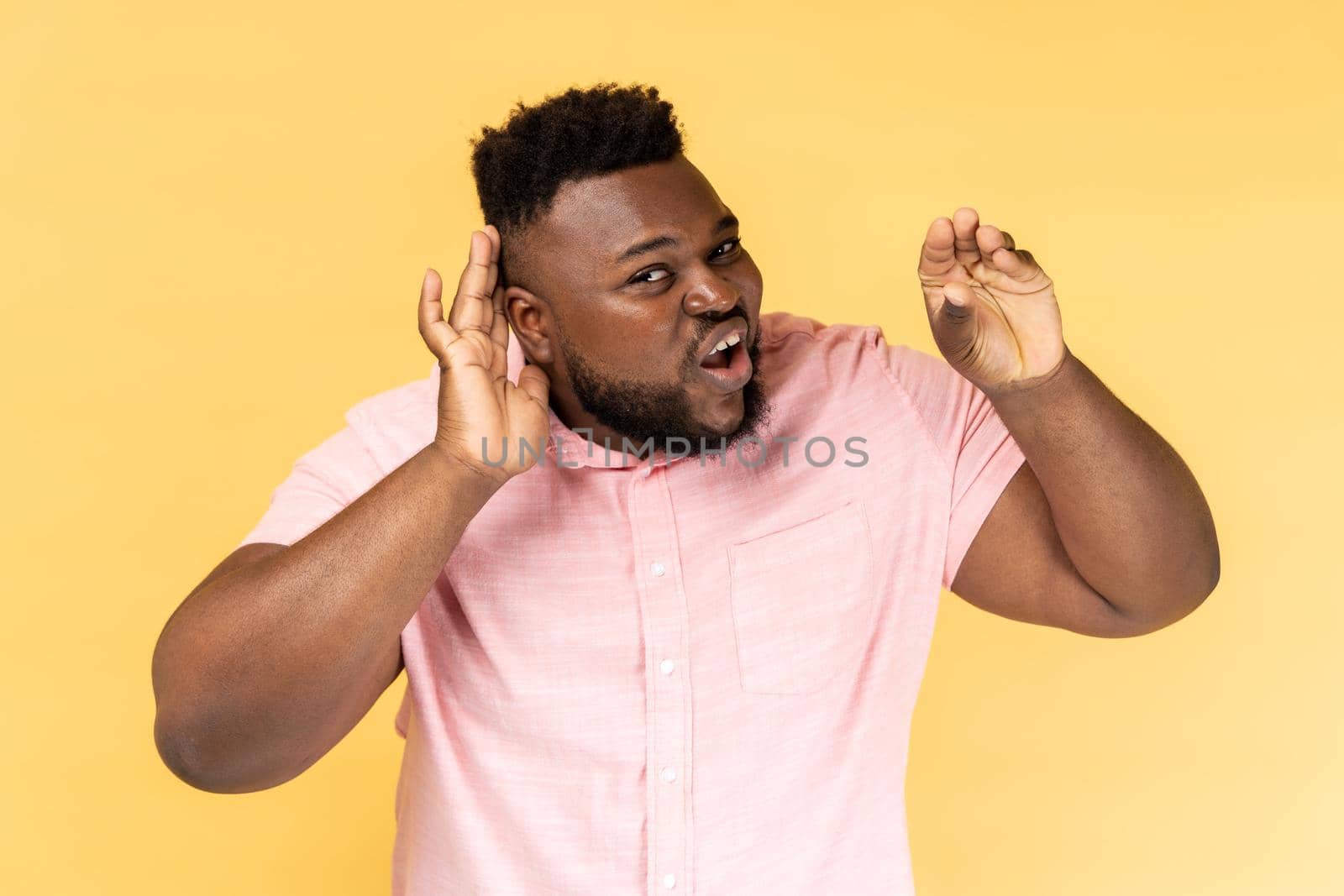 Portrait of unsure bearded young adult man wearing pink shirt standing with hand near ear, listening with frowning face, showing blah blah gesture. Indoor studio shot isolated on yellow background.