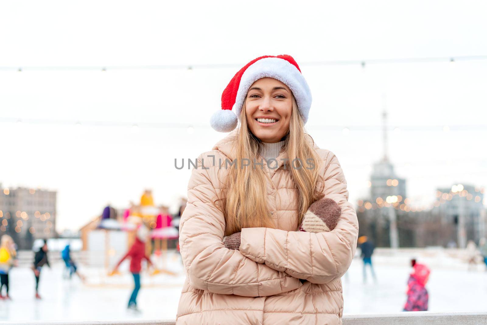 Outdoor portrait young beautiful fashionable woman wearing Santa hat, posing street of European city. Winter Christmas holidays concept. ice skate rink background. Caucasian female winter clothes