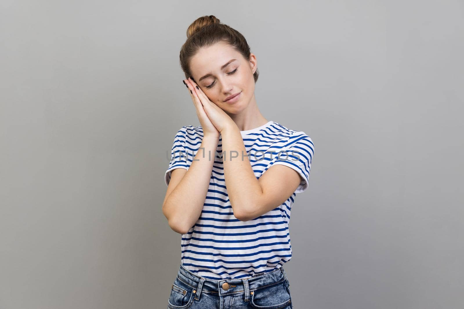 Portrait of sleepy bored woman wearing striped T-shirt leaning head on hands, keeps eyes closed, having nap, feeling tired and exhausted. Indoor studio shot isolated on gray background.
