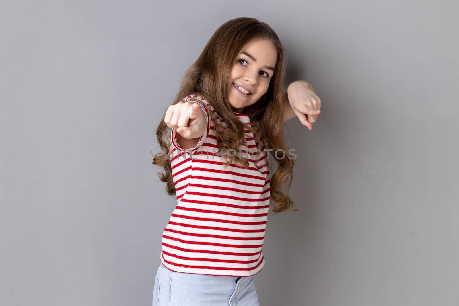 Portrait of joyful cheerful little girl wearing striped T-shirt pointing to camera, smiling, making happy choice, we need you concept. Indoor studio shot isolated on gray background.