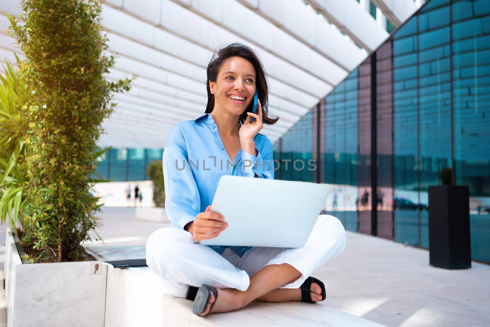 Businesswoman use laptop outdoor sitting near office building. Caucasian female business person 30 years have phone conversation and smile. Remote work freelancer concept. Freelance job its new normal