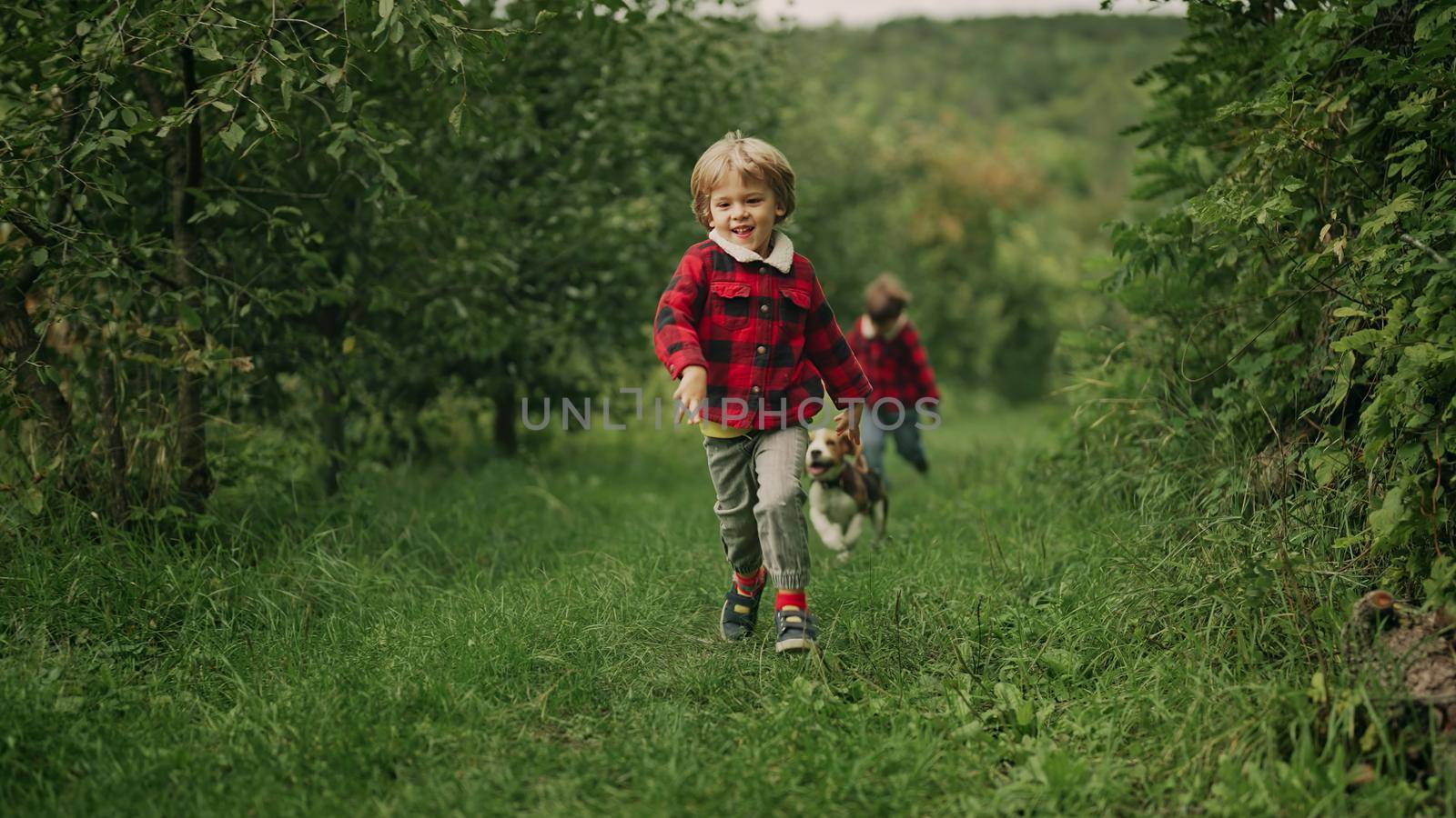 Happy kids running together with beagle puppy in green garden. Smiling brothers, stylish boys having fun with dog, playing outdoors. Friendship, family, twins, childhood concept. High 4k footage