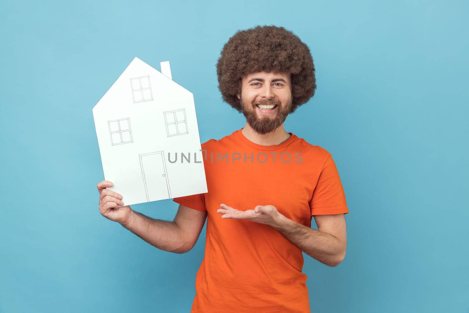 Man with Afro hairstyle presenting paper house with drawn windows and doors, making project of home of her dreams, looking at camera with smile. Indoor studio shot isolated on blue background.