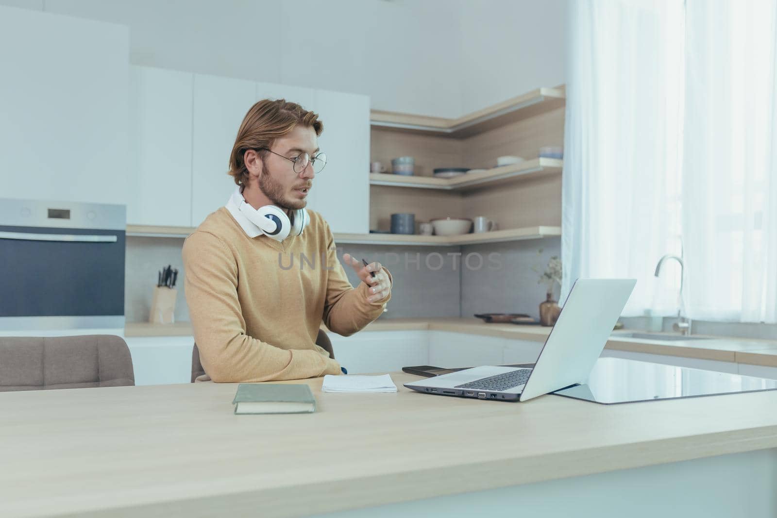 Young businessman working remotely from home sitting in kitchen, man using laptop for video call concentrating and talking seriously on online meeting with colleagues colleagues.