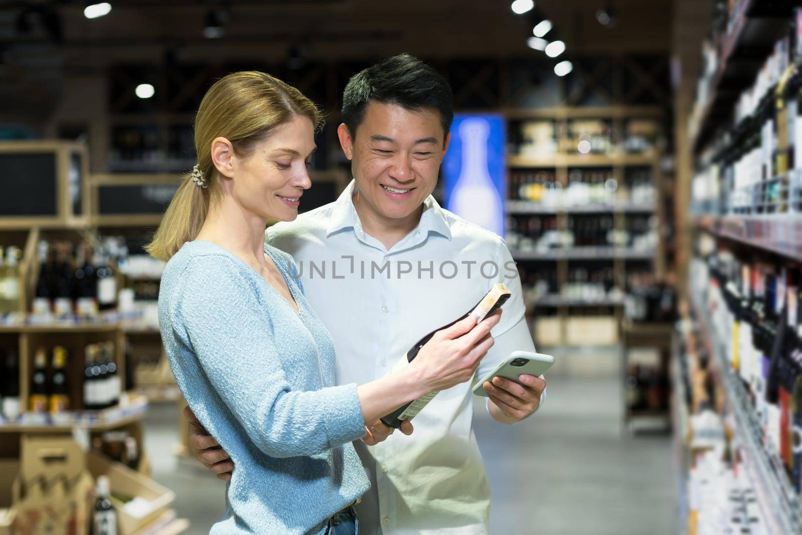 A young international family, an Asian man and a blonde woman, buy wine for dinner in the liquor department in a supermarket. They are holding a bottle, phone, satisfied with their choice.