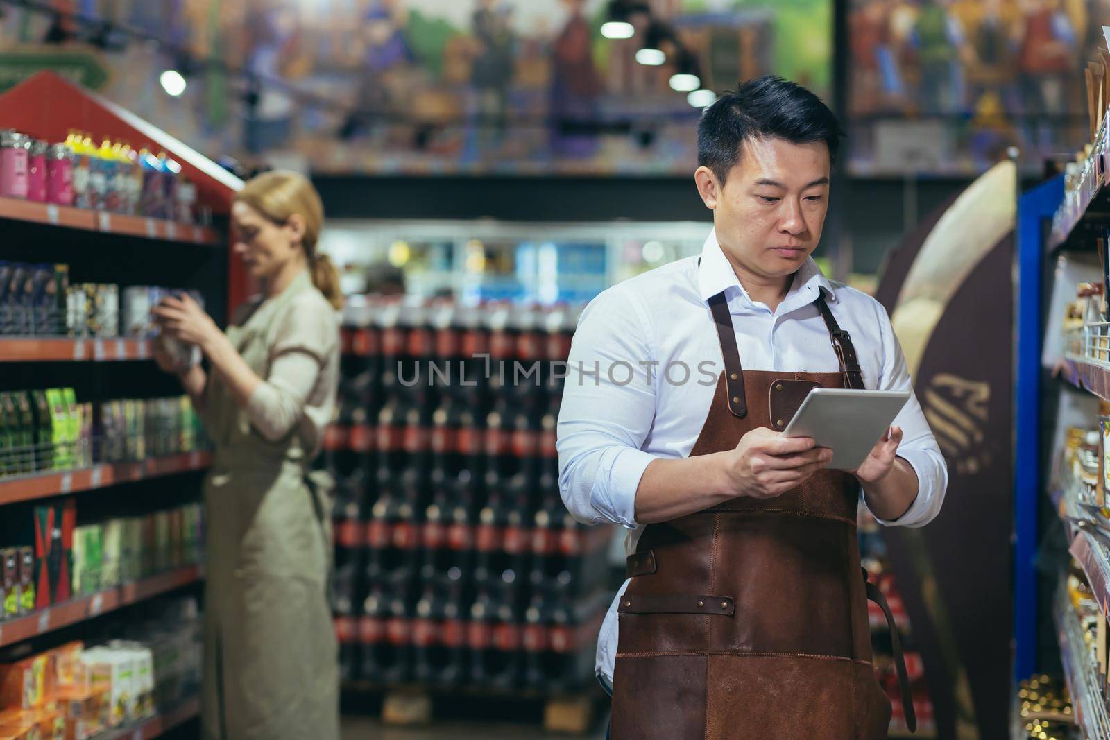 Portrait of young handsome Asian man, shop owner, supermarket worker. Standing in an apron near the shelves with goods, writing in a notebook. Female employee in the background.