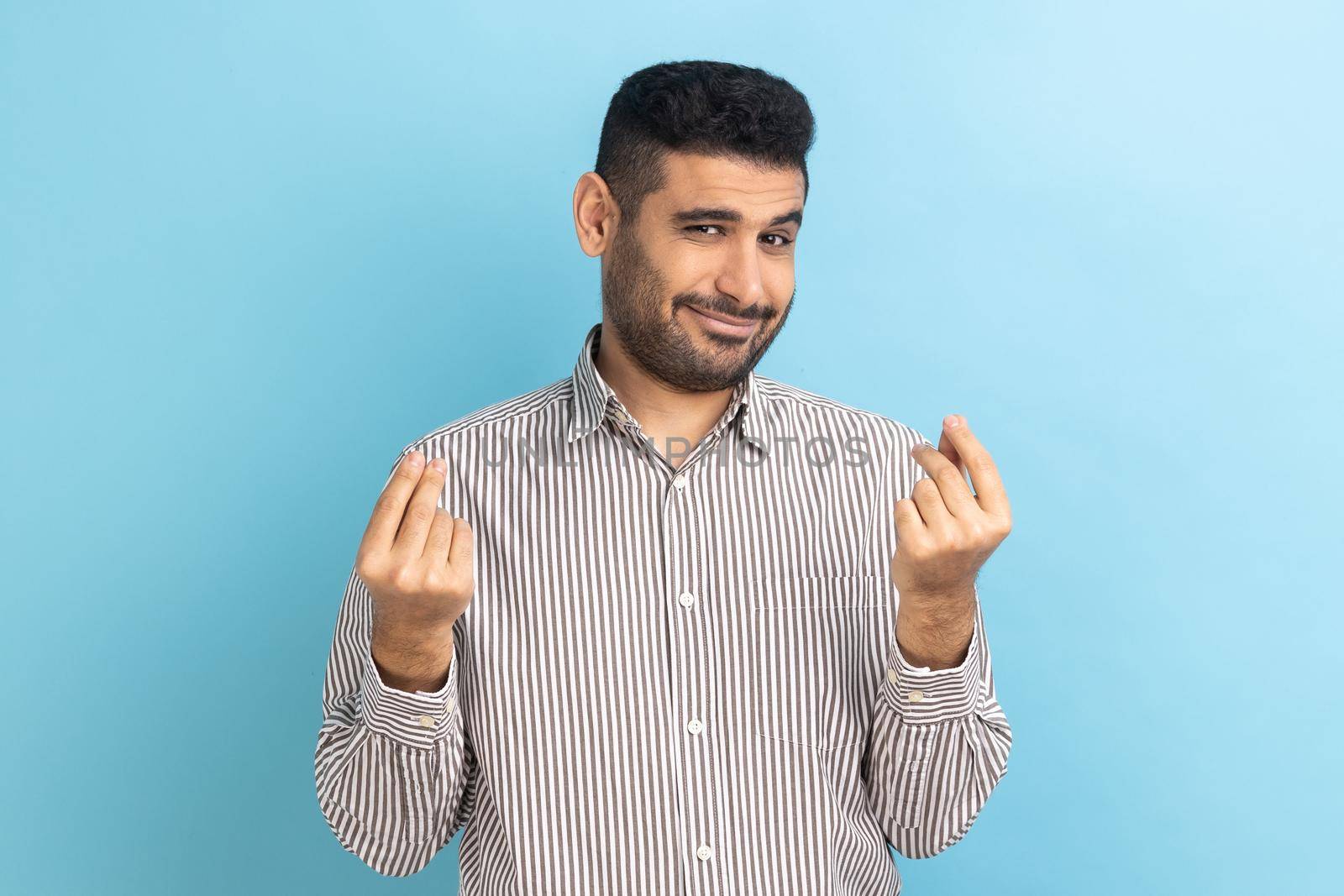 Cunning mercantile businessman with beard rubbing fingers showing money gesture, asking for salary, demanding bribe, wearing striped shirt. Indoor studio shot isolated on blue background.