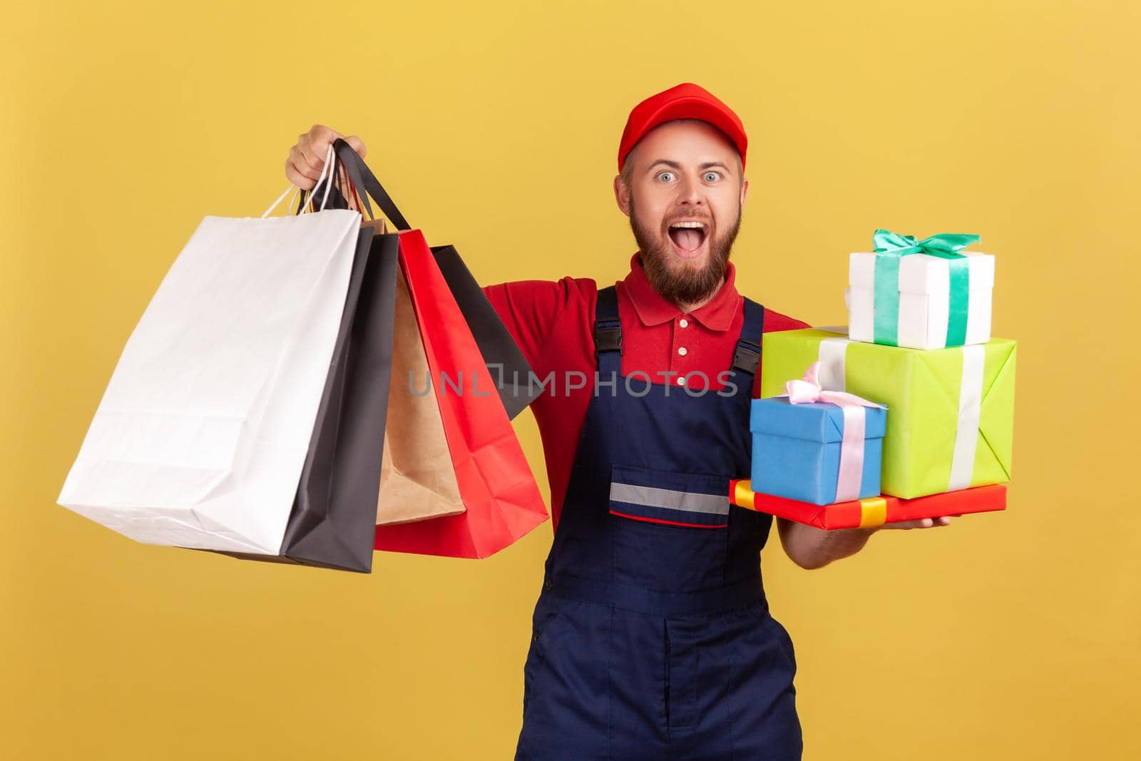 Portrait of excited amazed bearded courier man standing with many present boxes and shopping bags, door to door delivery, looking at camera. Indoor studio shot isolated on yellow background.