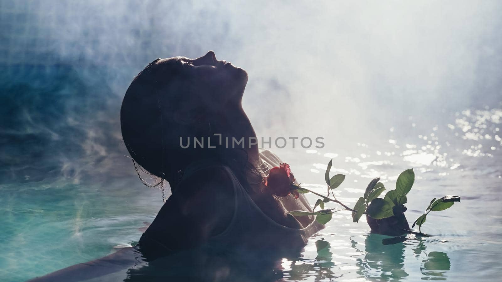 Silhouette of beautiful young woman stroking neck with rose flower in water on fog or smoke background. Sexy girl enjoying time in pool at night. High quality photo