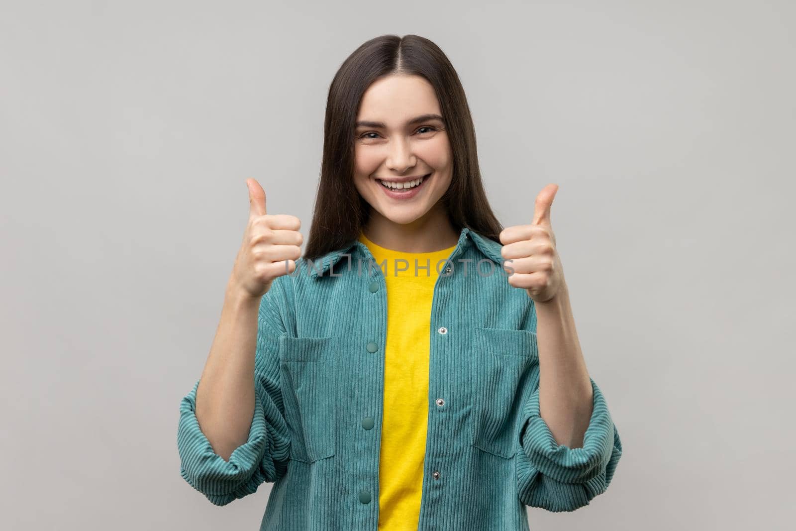 Happy woman looking at camera with toothy smile and showing thumbs up, approval sign, satisfied with service, good feedback, wearing casual style jacket. Indoor studio shot isolated on gray background