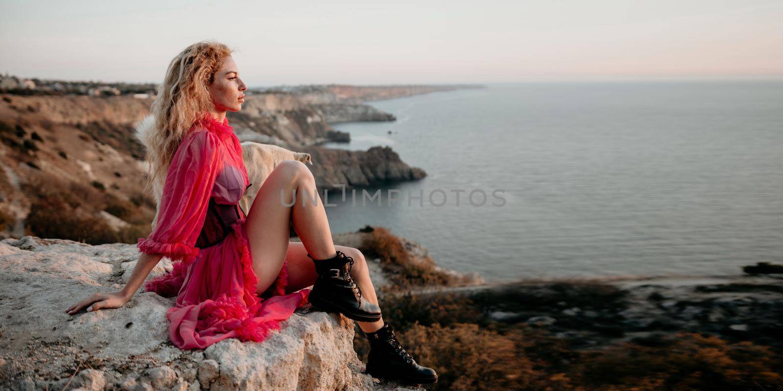 Close up portrait of curly redhead young caucasian woman with freckles looking at camera and smiling. Cute woman portrait in a pink long dress posing on a volcanic rock high above the sea at sunset by panophotograph