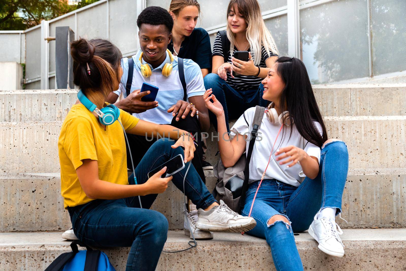 Happy multiracial teenage high school students sit on steps talking and using mobile phones outside. by Hoverstock