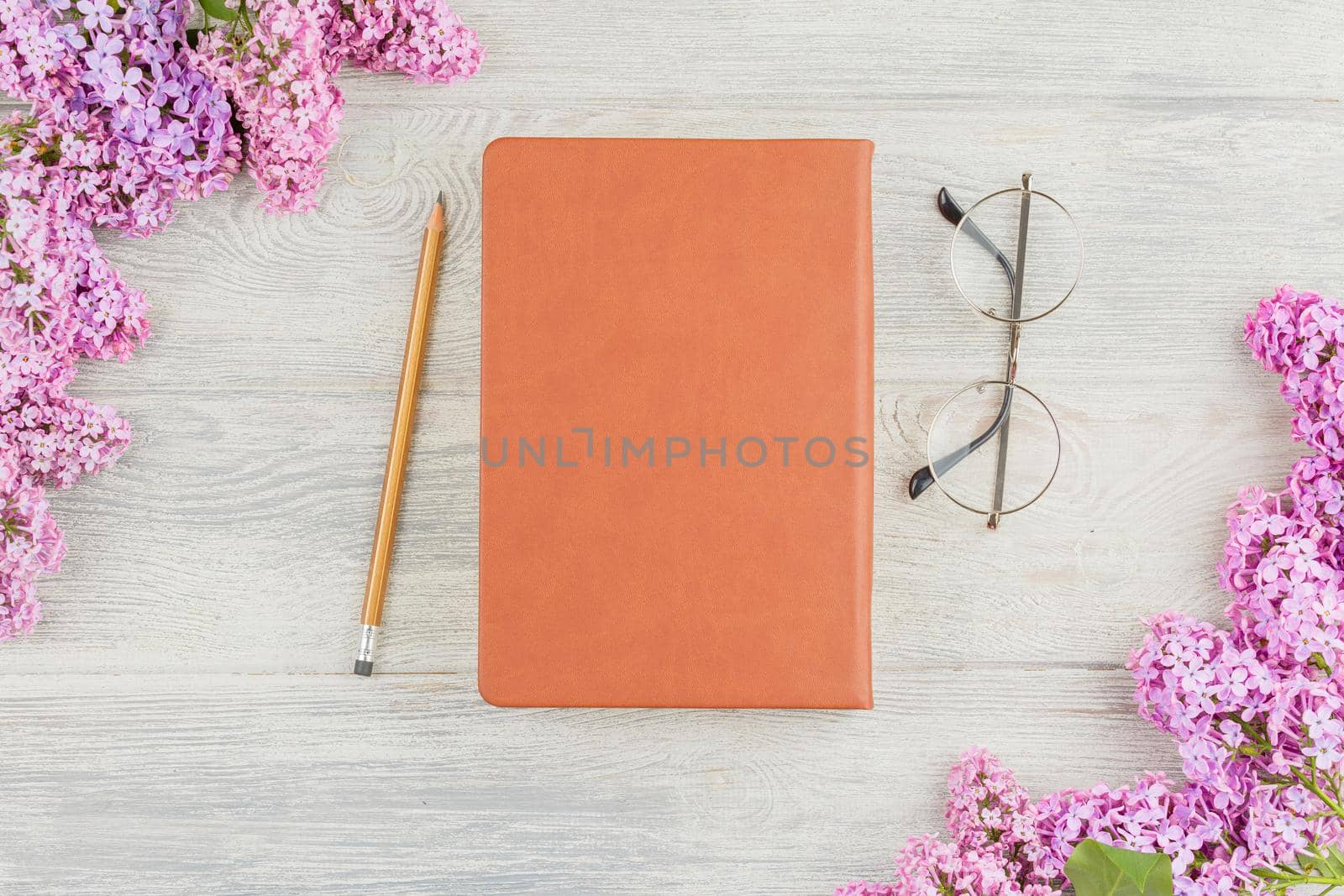 Leather book, pencil, glasses and lilac flowers on a white wooden background. Top view.