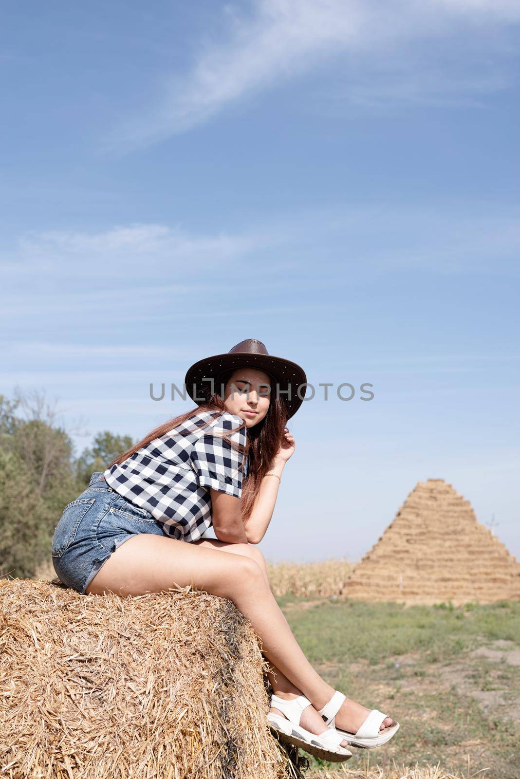 beautiful woman in plaid shirt and cowboy hat resting on haystack against sky background by Desperada