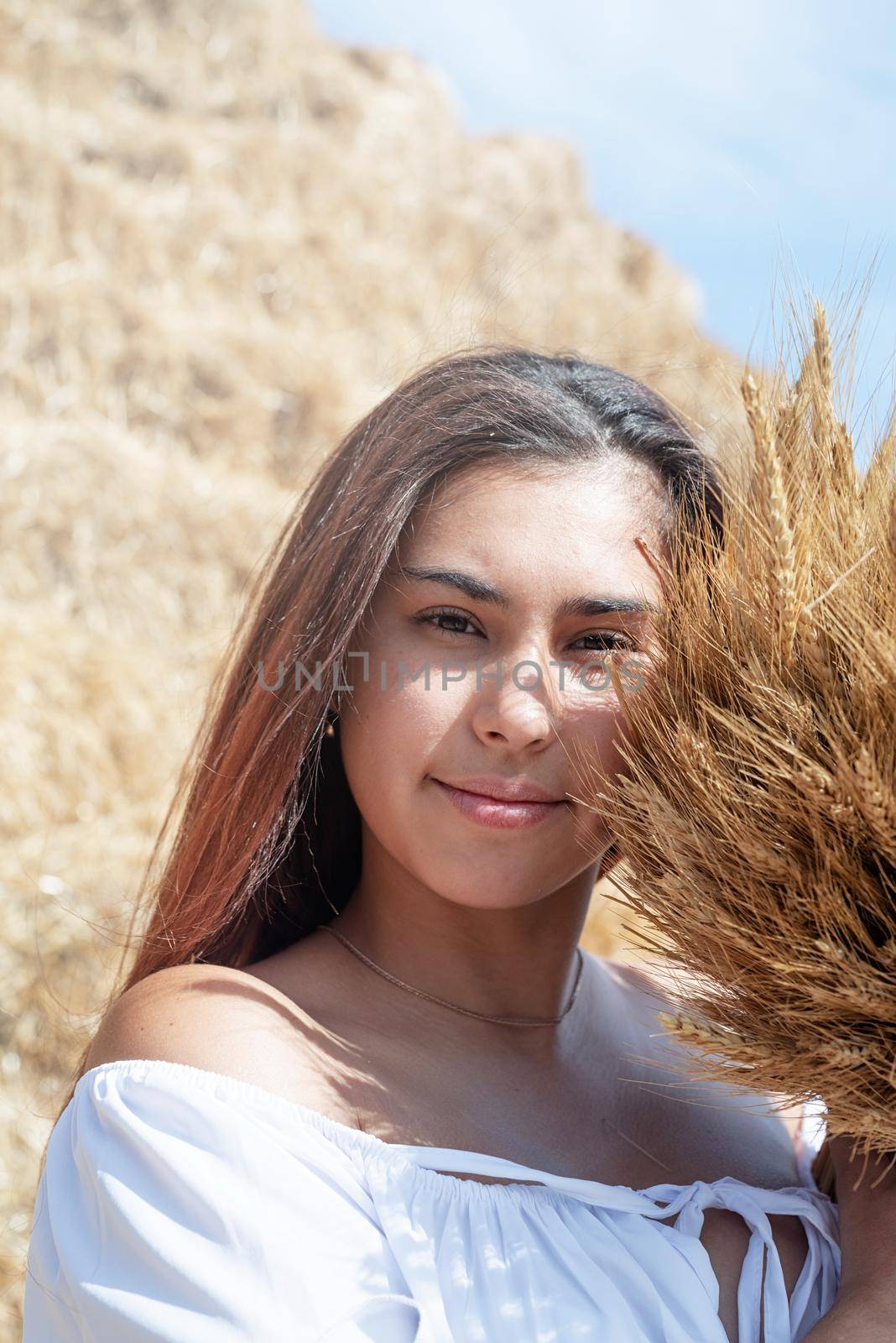 Picnic in countryside. Young woman sitting on haystack in harvested field holding wheat bouquet