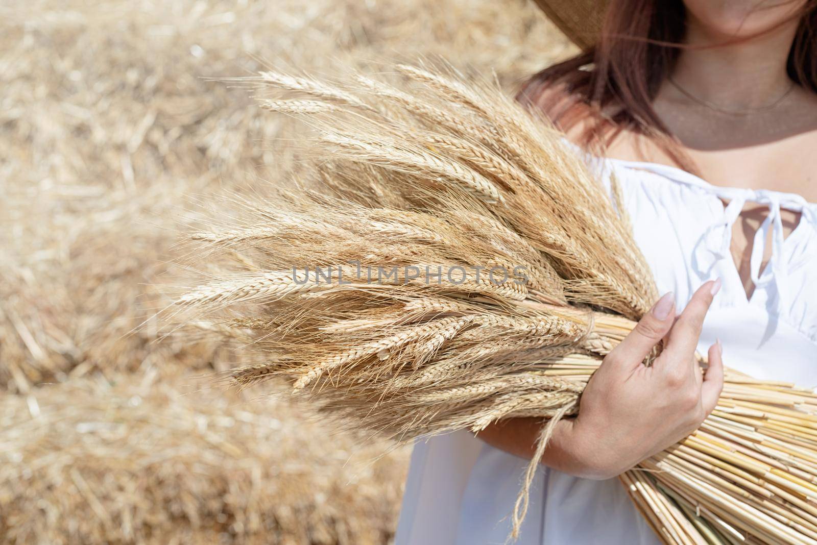 Picnic in countryside. Young woman sitting on haystack in harvested field holding wheat bouquet