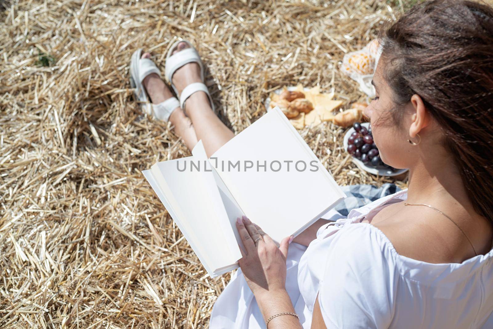 Picnic in countryside. Young woman in white dress sitting on haystack in harvested field, reading blank book. Book mockup