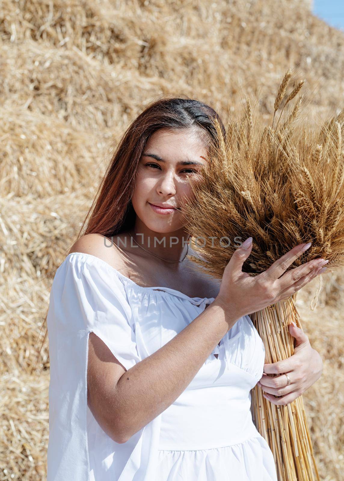 Young woman sitting on haystack in harvested field holding wheat bouquet by Desperada
