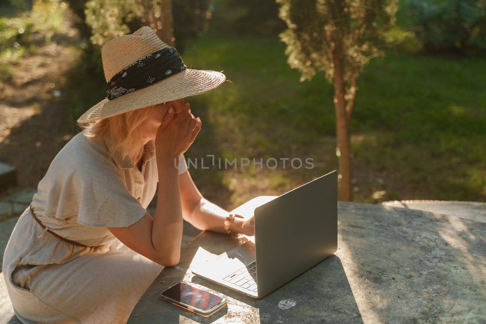 a beautiful mature woman in hat works on a computer at a white table in nature and spends her day productively by Matiunina