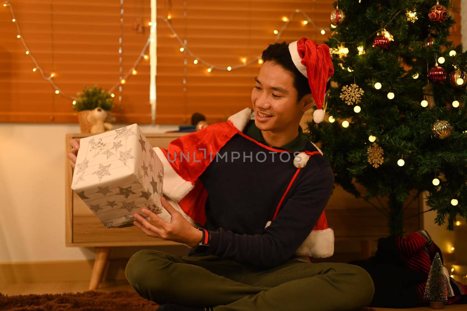 Man in Santa hat opening Christmas gifts, sitting near Christmas tree in decorated room lighted with soft lights.