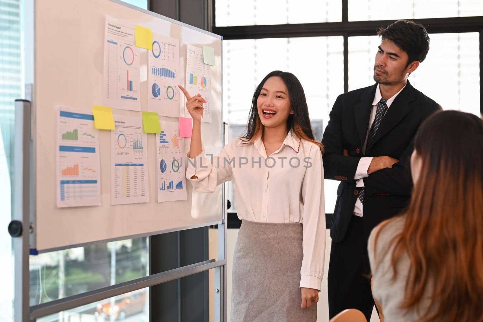Confident young woman standing near flip chart with her colleague and giving presenting new marketing strategy corporate meeting.