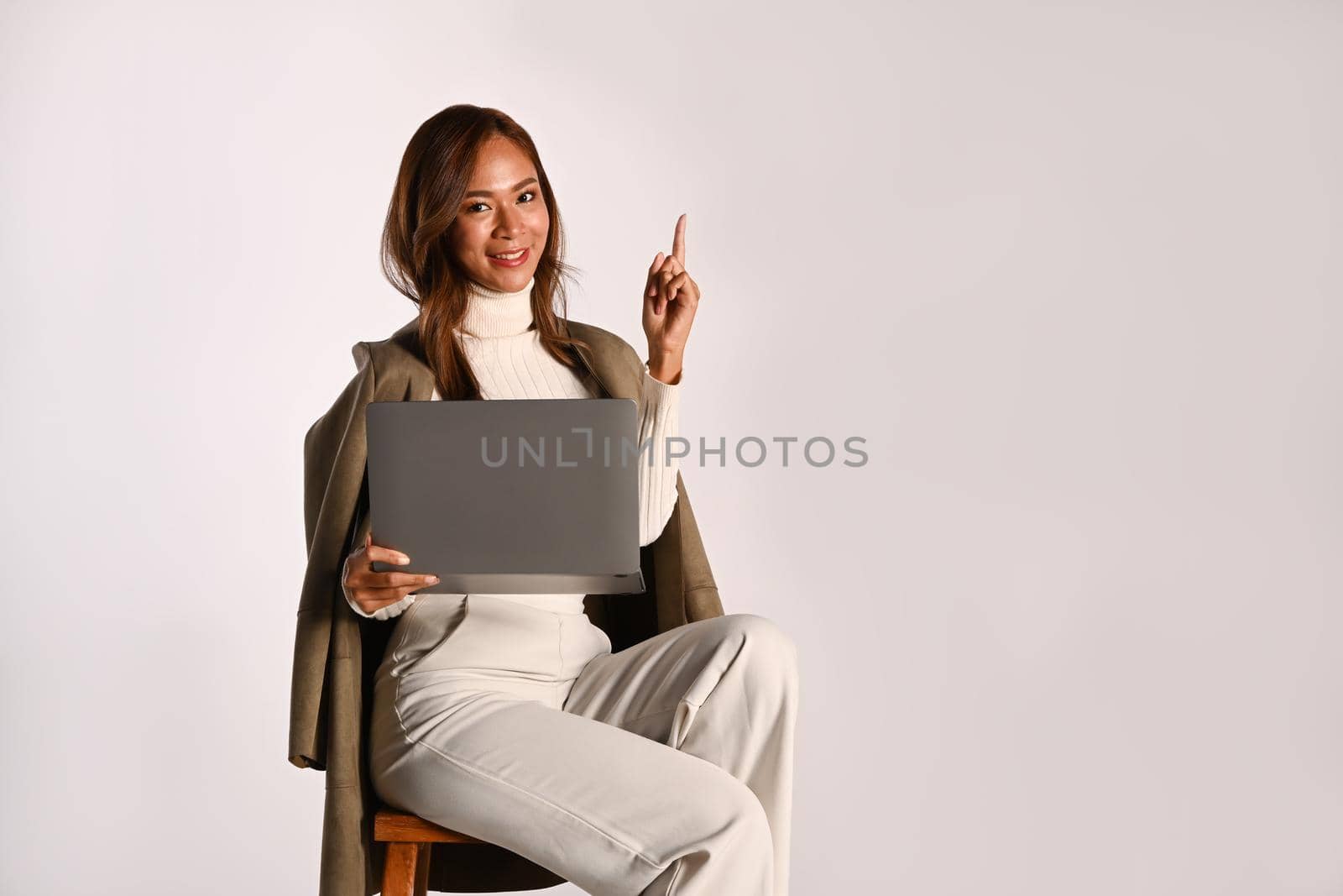 Gorgeous young woman sitting with laptop and pointing at copy space over white studio background. People and technology concept.
