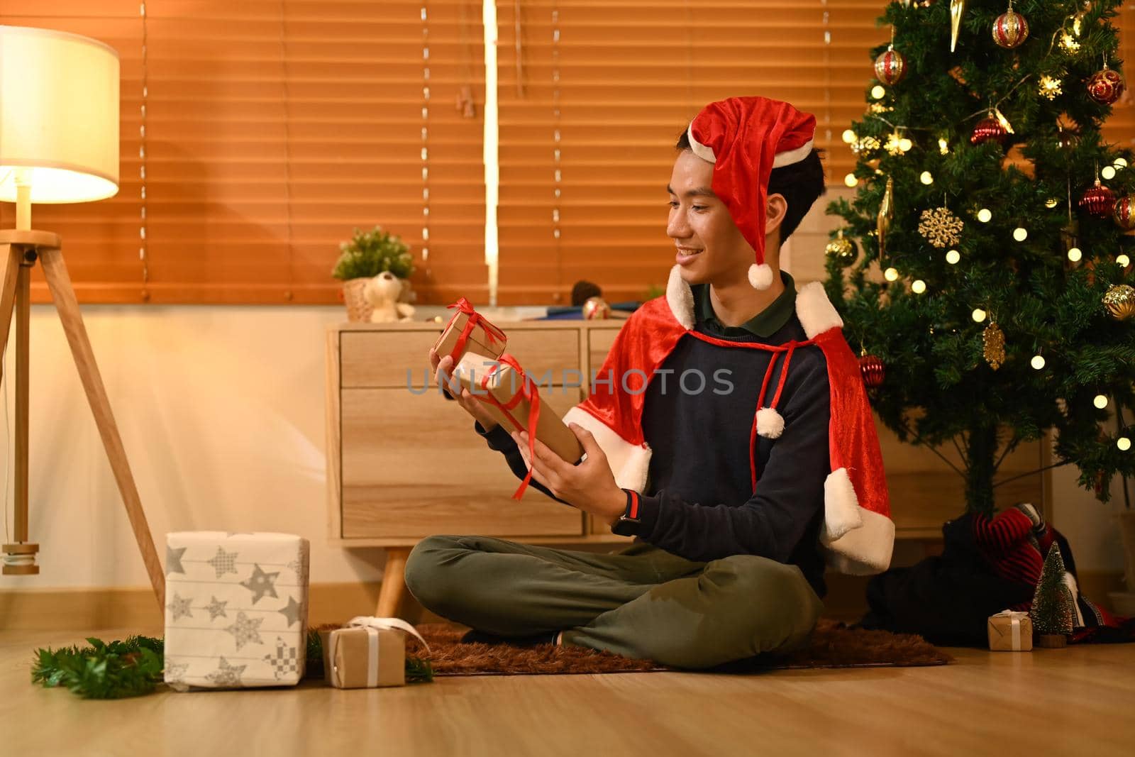 Happy man preparing Christmas presents while sitting near decorated Christmas tree in living room lighted with soft lights.