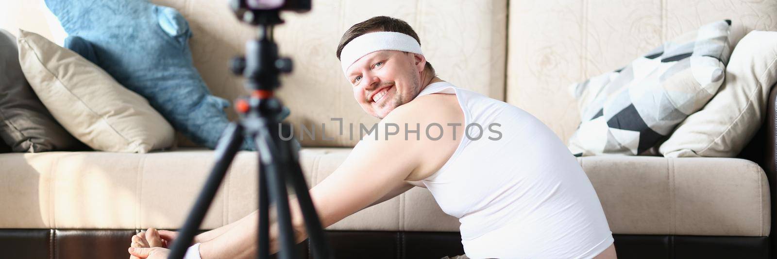 Low angle of happy man in sportswear exercise with joy, stretching on floor carpet. Male filming video for channel on social media. Internet, sport concept