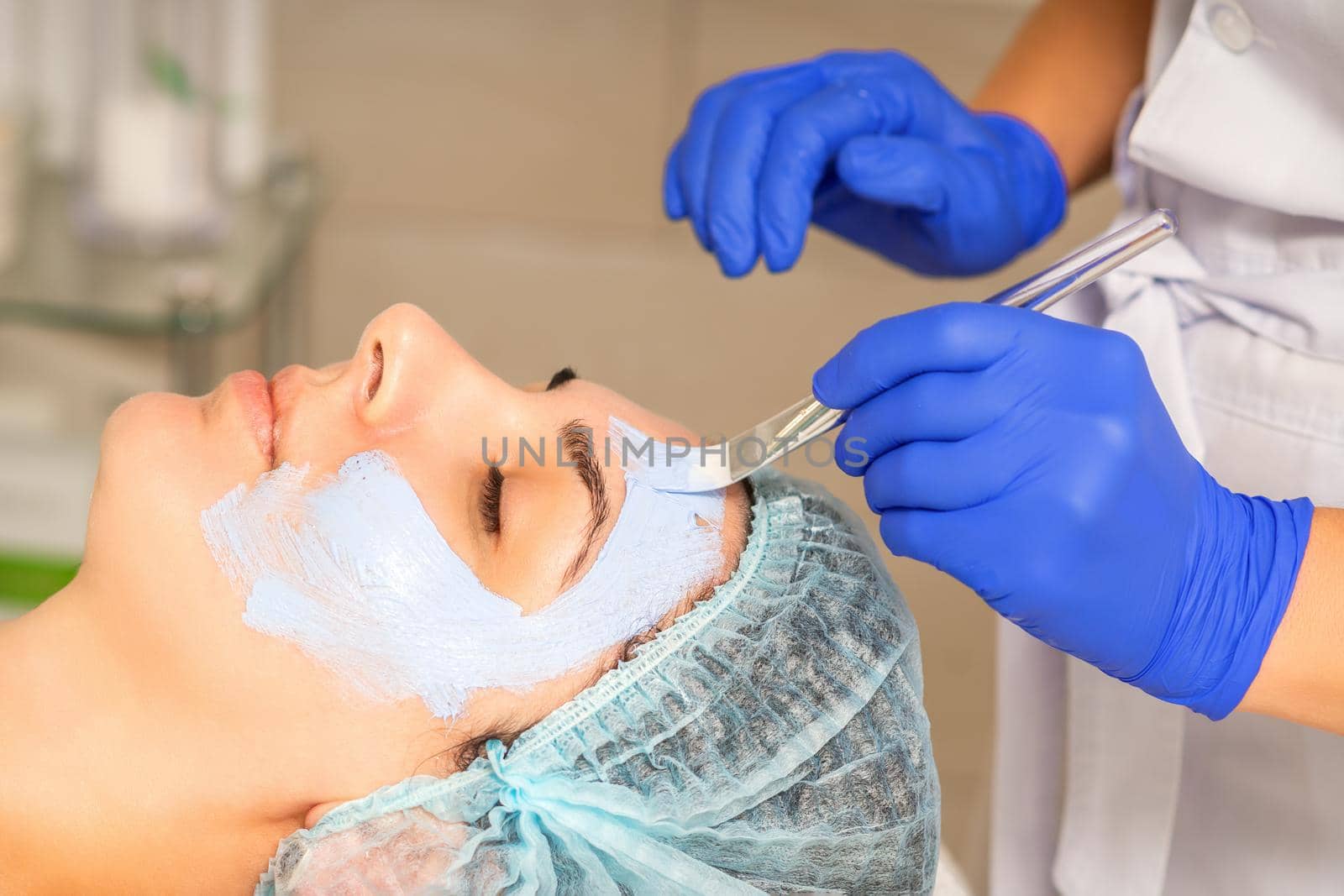 The beautician with brush applies a photochemical and glycolic peeling face mask to the female patient face in the beauty clinic