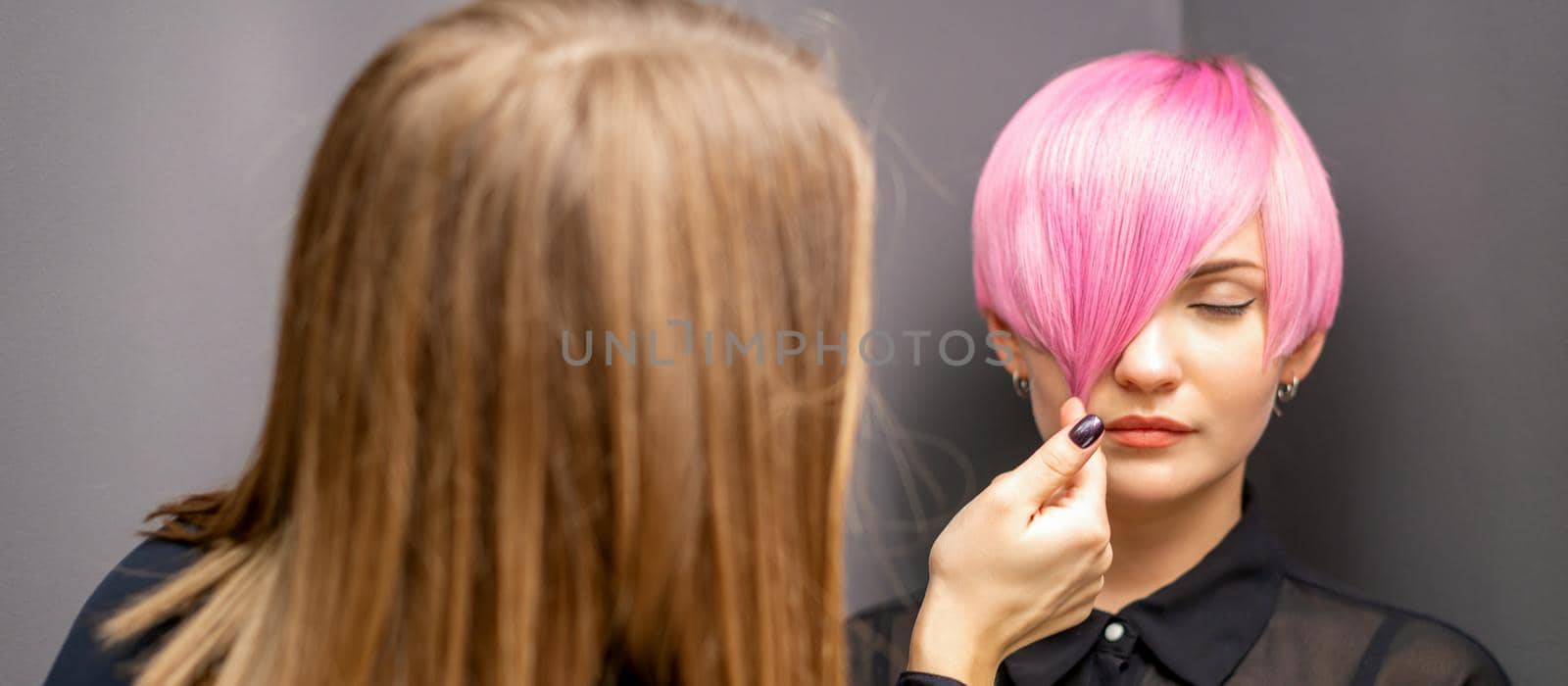 Hairdresser with hands is checking out and fixing the short pink hairstyle of the young white woman in a hair salon