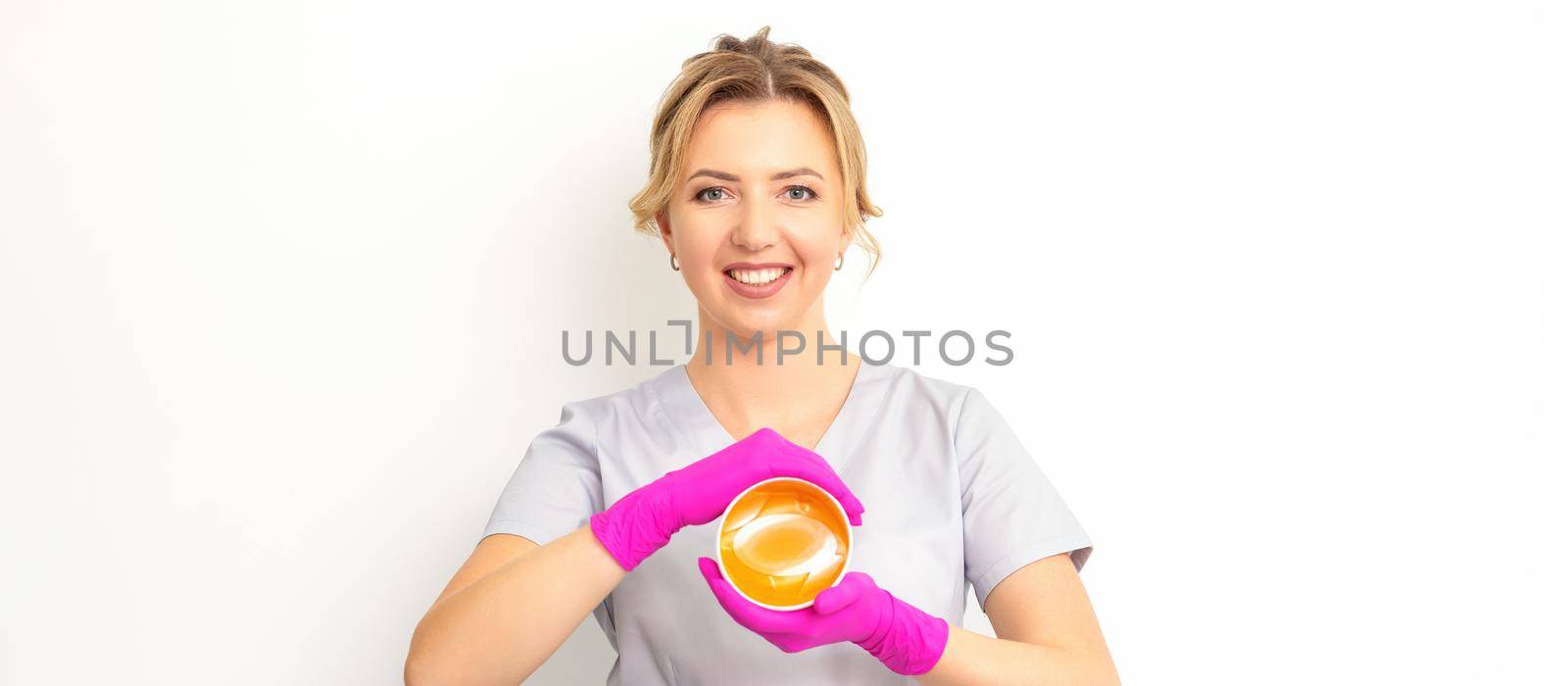 Portrait of a female caucasian beautician holding a jar of sugar paste for sugaring wearing pink gloves on white background. by okskukuruza