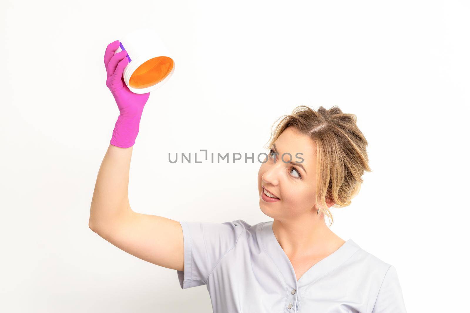 Portrait of a female caucasian beautician holding a jar of sugar paste for sugaring wearing pink gloves on white background