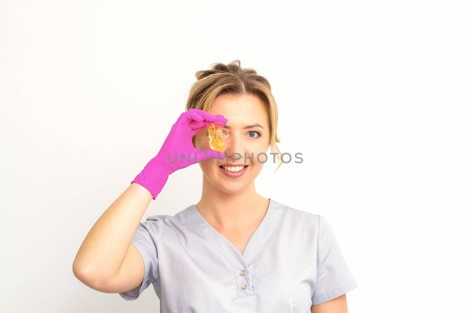 The master of sugar hair removal holds liquid yellow sugar paste, covering her eye with wax for depilation on a white background. by okskukuruza