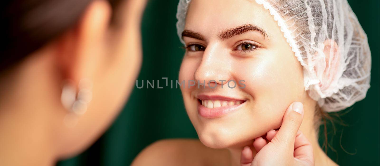 Plastic surgery and beauty concept. Professional cosmetician examining and touching the face of the beautiful young caucasian smiling woman in a clinic of esthetic cosmetology. by okskukuruza