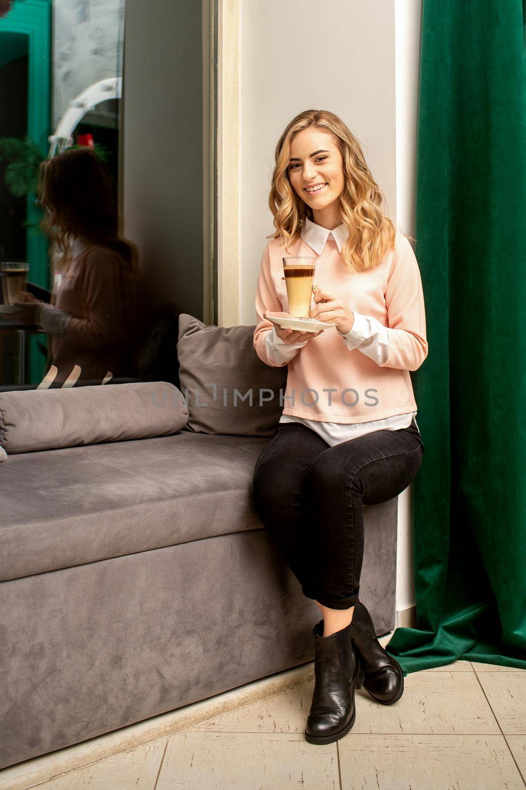Beautiful caucasian young smiling woman with long wavy blonde hair holding and drinking a latte from a glass cup, sitting on a sofa. by okskukuruza
