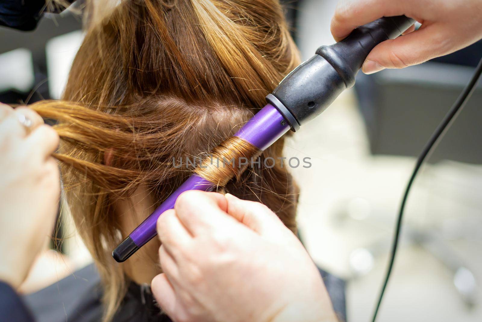 The hairstylist makes curls hairstyle of long brown hair with the curling iron in hairdresser salon, close up. by okskukuruza