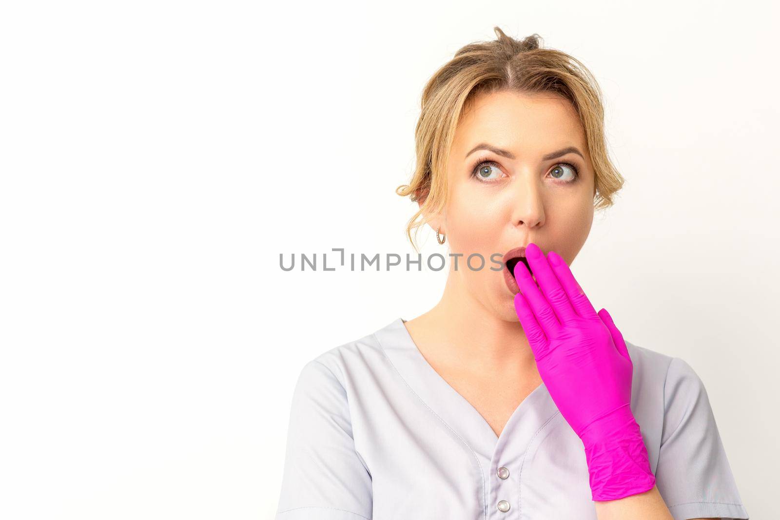 Portrait of a young female caucasian doctor or nurse is shocked covering her mouth with her pink gloved hands against a white background. by okskukuruza